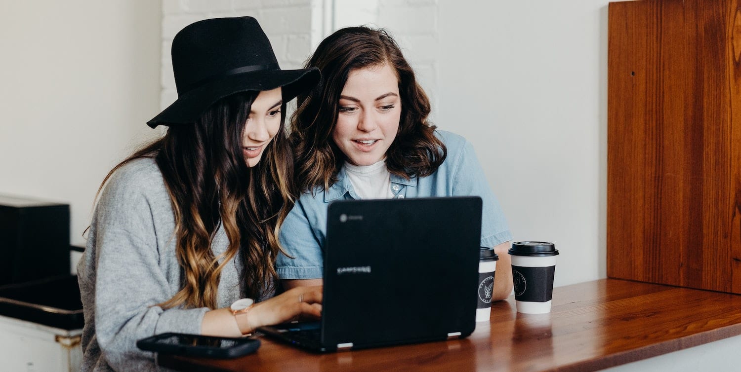 Image of two young women sitting at a desk with a laptop and chatting.