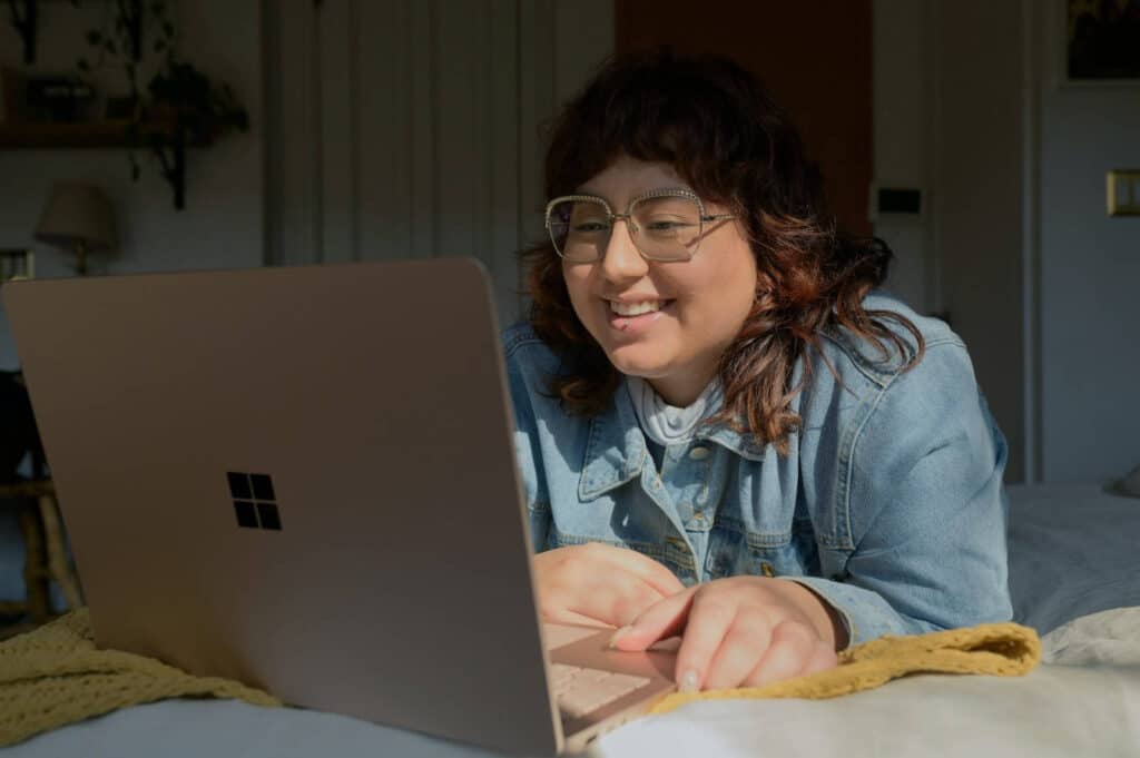 a woman wearing glasses using her laptop on her bed