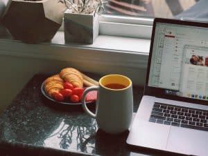 Image of a laptop on a desk. Next to it there is a cup of coffee and some food.