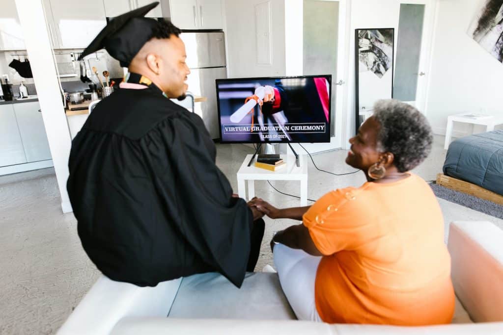 Image of a graduating student and his family member chatting and watching a graduation ceremony on their TV.